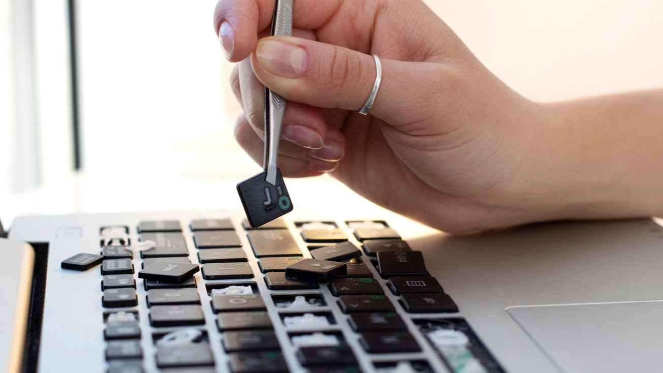 A woman's hand holding a key with tweezers above a laptop keyboard with numerous missing keys.