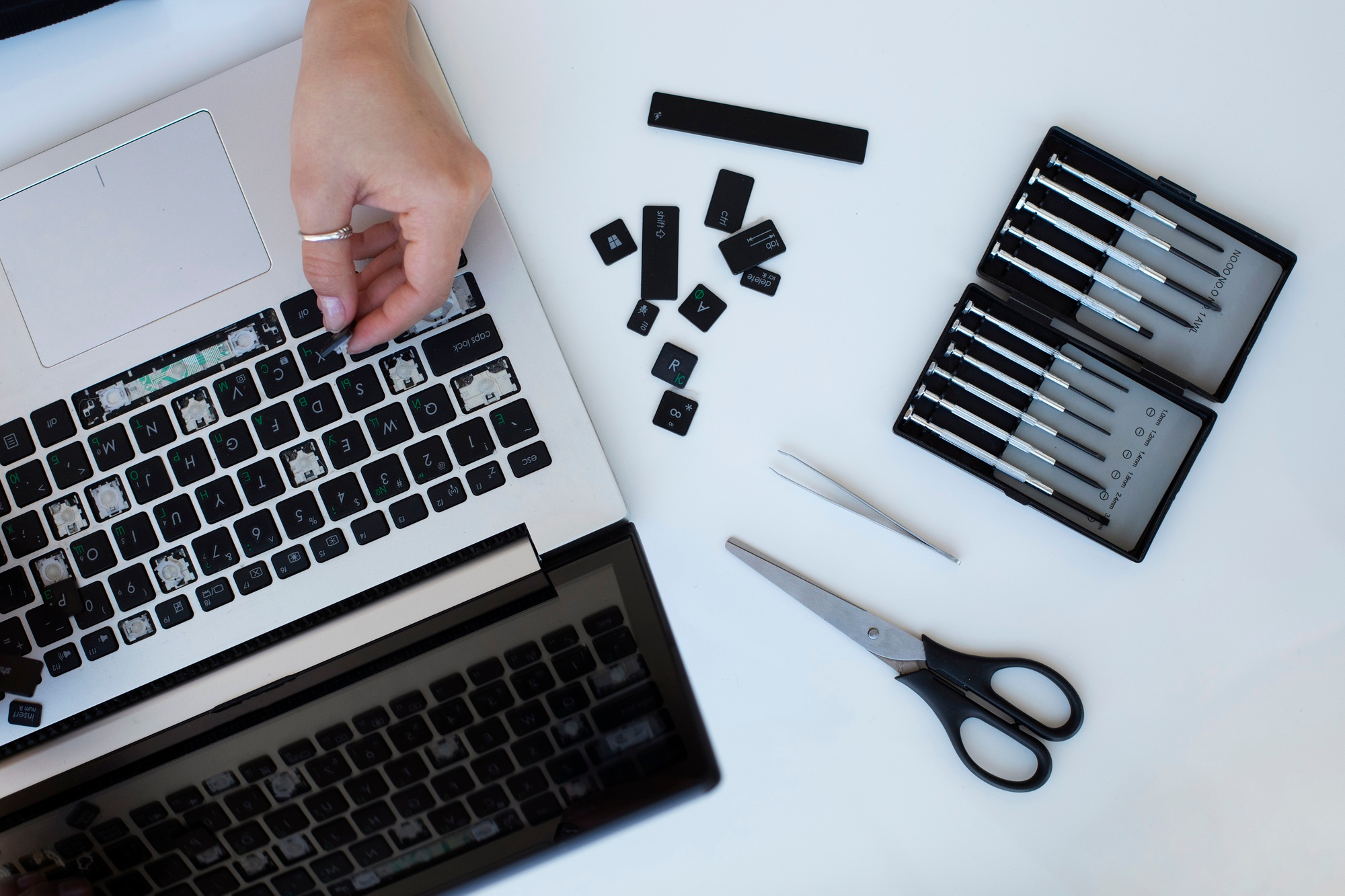 A laptop keyboard with removed keys, tools, and a hand of a person working on its repair.